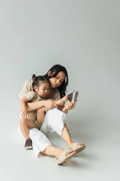 Full length of asian mother sitting and wearing shoe of foot of toddler daughter on grey — Stock Photo