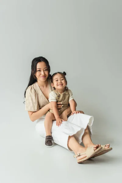 Full length of cheerful asian mother sitting with toddler daughter on grey — Stock Photo