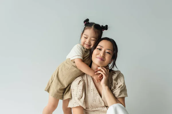 Fröhlich asiatisch kleinkind tochter umarmt glücklich mutter isolated auf grau — Stockfoto