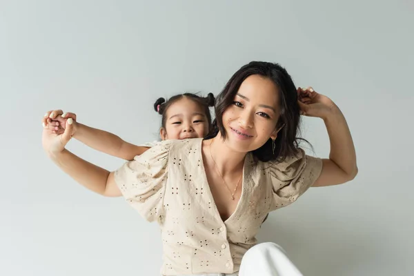 Cheerful asian woman holding hands with toddler daughter isolated on gray — Stock Photo