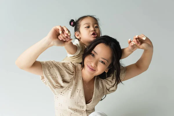 Улыбающаяся азиатка, держащаяся за руки с дочкой малыша, изолированной на сером — стоковое фото