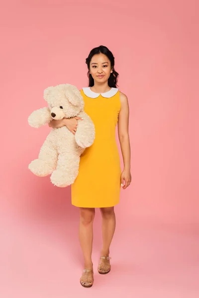 Full length of happy asian young woman in yellow dress holding teddy bear on pink — Stock Photo