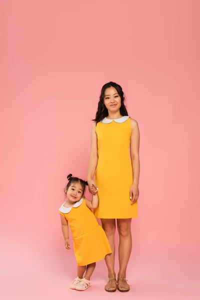 Pleased asian woman holding hands with smiling toddler daughter on pink — Stock Photo