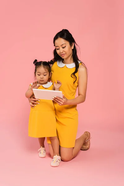 Asian mother holding digital tablet near toddler daughter on pink — Stock Photo