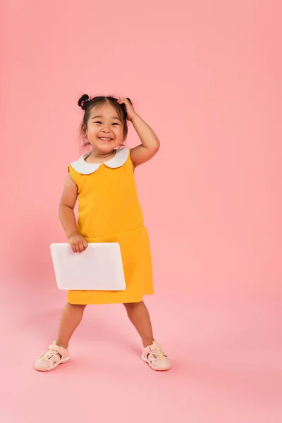 Full length of happy asian toddler kid in yellow dress holding digital tablet on pink — Stock Photo