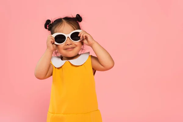 Alegre asiático niño pequeño en amarillo vestido ajuste gafas de sol aislado en rosa — Stock Photo