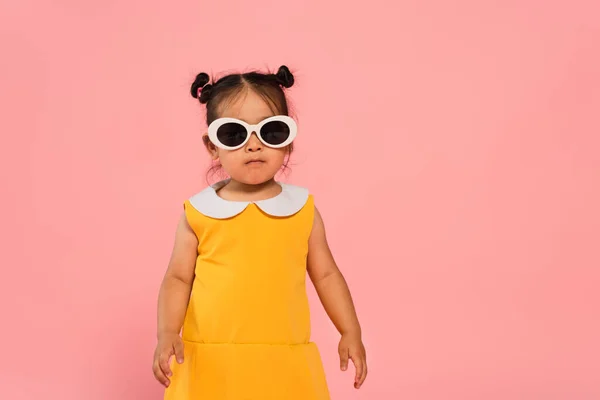 Asiático niño pequeño en amarillo vestido y gafas de sol posando aislado en rosa - foto de stock