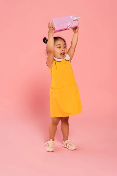 Full length of cheerful asian toddler kid in yellow dress holding present above head on pink — Stock Photo