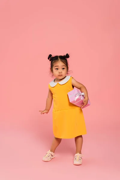 Full length of asian toddler kid in yellow dress holding present above head on pink — Stock Photo
