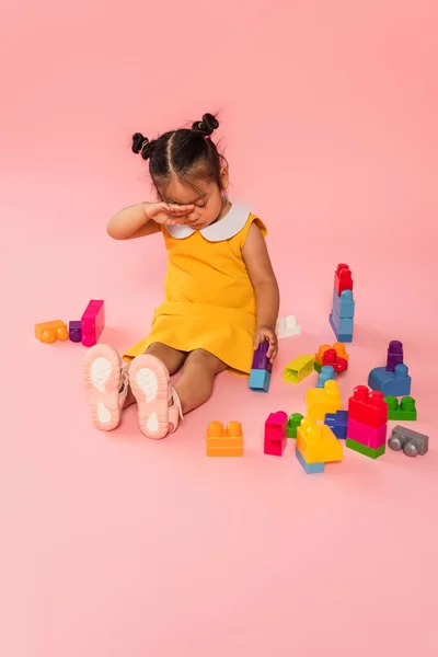 Tired asian toddler kid in yellow dress playing building blocks on pink — Stock Photo
