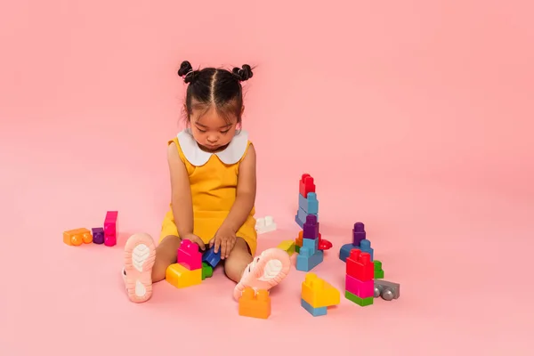 Asian toddler girl in yellow dress playing building blocks on pink — Stock Photo