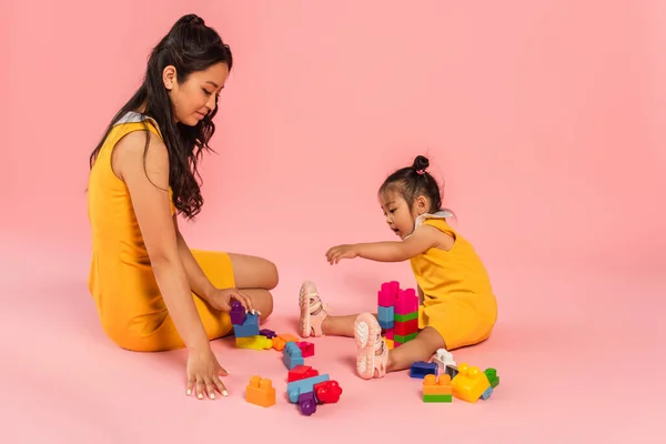 Азіатська мати і дочка, сидячи і граючи будівельні блоки на рожевому — стокове фото