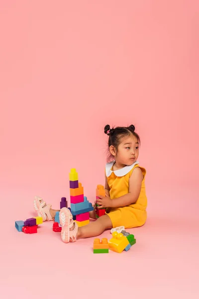 Distracted asian toddler girl in yellow dress playing building blocks on pink — Stock Photo