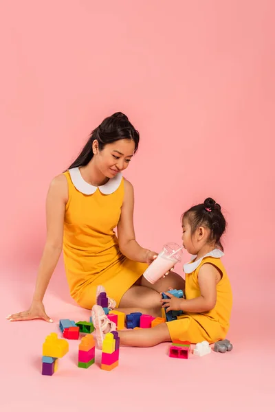 Feliz asiático mujer y hija sentado y jugando bloques de construcción en rosa - foto de stock