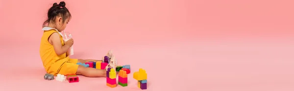 Asian toddler girl in yellow dress drinking milkshake through straw near colorful building blocks on pink, banner — Stock Photo