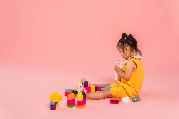 Menina asiática em vestido amarelo bebendo milkshake através de palha perto de blocos de construção coloridos em rosa — Fotografia de Stock