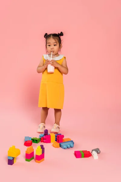Volle Länge der asiatischen Kleinkind Mädchen in gelbem Kleid trinken leckeren Milchshake durch Stroh in der Nähe von bunten Bausteinen auf rosa — Stockfoto