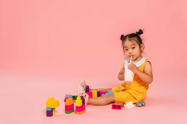 Asiático criança criança em vestido amarelo beber saboroso milkshake através de palha perto de blocos de construção coloridos em rosa — Fotografia de Stock