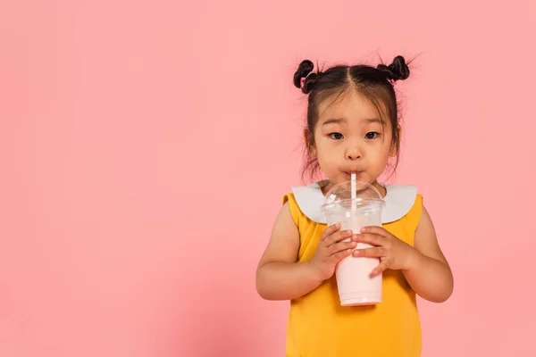 Азиатский малыш в жёлтом платье пьет вкусный молочный коктейль через соломинку на розовом — стоковое фото