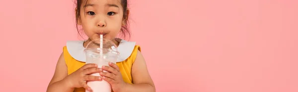 Asiatique tout-petit enfant en robe jaune boire savoureux milkshake à travers la paille isolé sur rose, bannière — Photo de stock