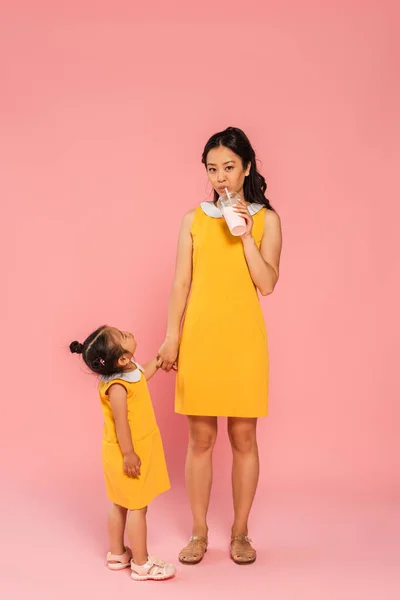 Азиатский малыш в жёлтом платье смотрит на мать, пьющую вкусный молочный коктейль через соломинку на розовом — стоковое фото