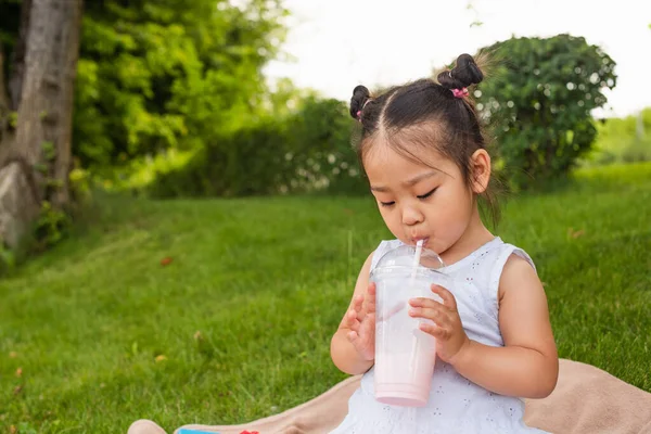 Азиатский малыш в платье пьет вкусный молочный коктейль через соломинку в парке — стоковое фото