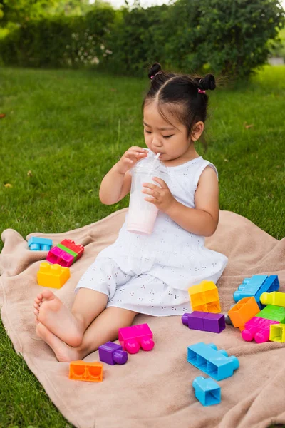 Asiático niño chica en vestido beber sabroso milkshake cerca construcción bloques en picnic manta - foto de stock