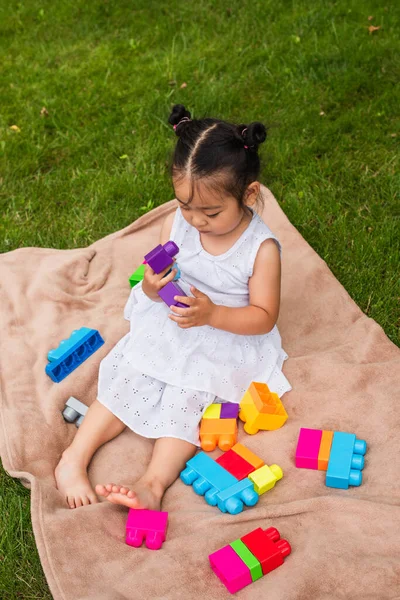 Високий кут зору на азіатську дитину в сукні, що грає будівельні блоки на пікніку ковдру в парку — стокове фото