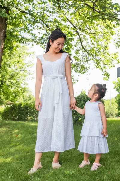 Longitud completa de feliz madre y sonriente niño asiático en vestidos tomados de la mano en parque - foto de stock