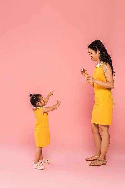 Full length of asian mother looking at toddler daughter pointing at soap bubble on pink — Stock Photo