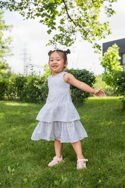 Comprimento total de alegre asiático menina no vestido de pé na grama verde — Fotografia de Stock