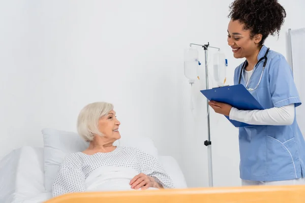 Sourire infirmière afro-américaine avec presse-papiers regardant le patient âgé — Photo de stock