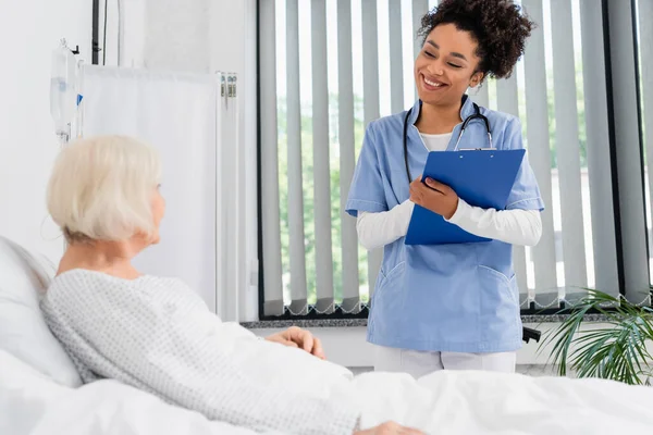 Enthousiaste infirmière afro-américaine avec presse-papiers regardant le patient âgé sur le lit — Photo de stock