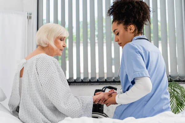 Seitenansicht einer afrikanisch-amerikanischen Krankenschwester, die auf einer Krankenhausstation den Puls einer älteren Frau überprüft — Stockfoto