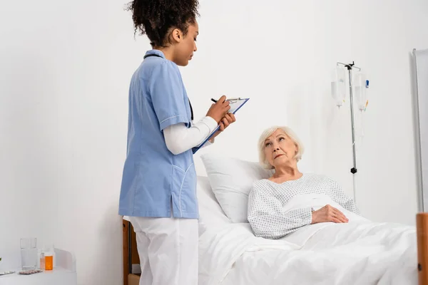 Enfermera afroamericana escribiendo en portapapeles cerca de paciente anciano en cama de hospital - foto de stock