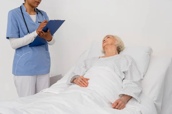 African american nurse writing on clipboard near senior patient lying on hospital bed — Stock Photo