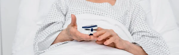 Cropped view of elderly patient holding oximeter on bed in clinic, banner — Stock Photo