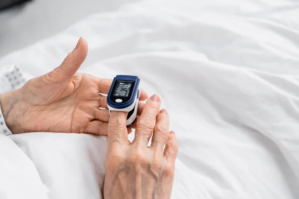 Обрізаний вид на пацієнта похилого віку, який використовує цифровий оксиметр на лікарняному ліжку — стокове фото