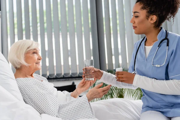 Vista lateral de una enfermera afroamericana sosteniendo un vaso de agua y pastillas cerca de un paciente mayor sonriente - foto de stock