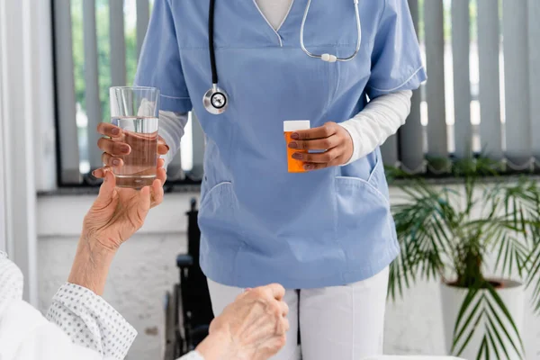 Обрізаний вид афроамериканської медсестри, що тримає таблетки і склянку води біля старшого пацієнта — стокове фото