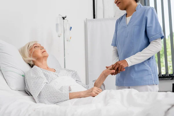 Африканская американская медсестра держит за руку пожилого пациента с носовым канюлем на больничной койке — стоковое фото