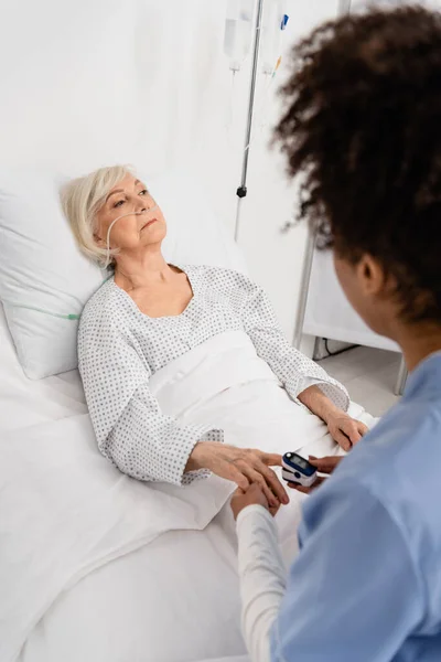 Patiente âgée avec canule nasale couchée sur le lit près d'une infirmière afro-américaine tenant un oxymètre en clinique — Photo de stock