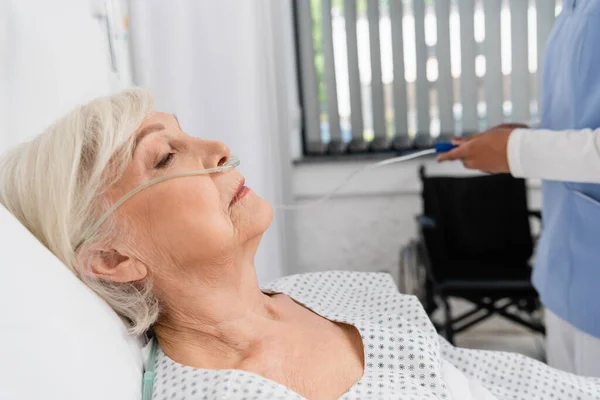 Visão lateral da mulher idosa com cânula nasal perto de enfermeira afro-americana turva no hospital — Fotografia de Stock