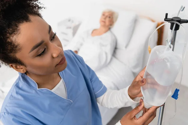 High angle view of african american nurse holding intravenous therapy bottle near blurred patient — Stock Photo