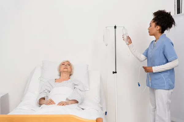 African american nurse holding bottle of intravenous therapy near senior patient in hospital — Stock Photo