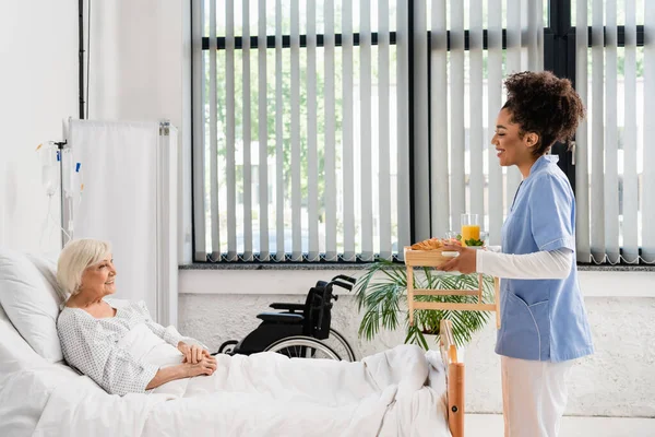Side view of smiling african american nurse holding food on tray near senior patient — Stock Photo