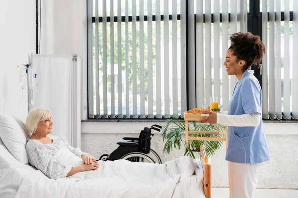 Seitenansicht einer lächelnden afrikanisch-amerikanischen Krankenschwester, die in der Nähe eines älteren Patienten Lebensmittel auf Tablett hält — Stockfoto