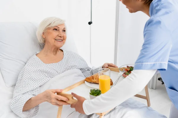 Sorridente paziente anziano guardando sfocata infermiera africana americana con il cibo in ospedale — Foto stock