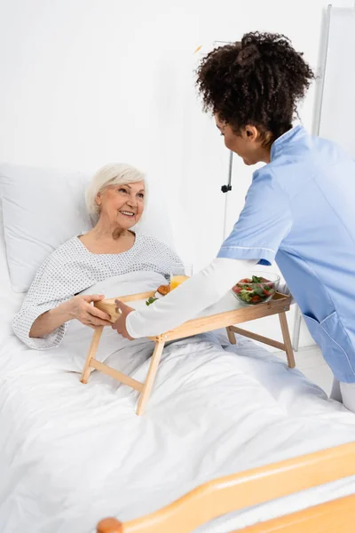 African american nurse holding tray with food near cheerful patient on bed in hospital ward — Stock Photo