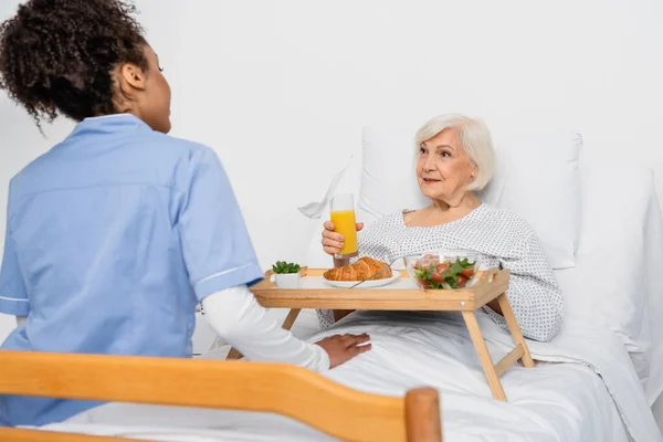 Patiente âgée regardant une infirmière afro-américaine près de la nourriture sur un plateau sur un lit d'hôpital — Photo de stock